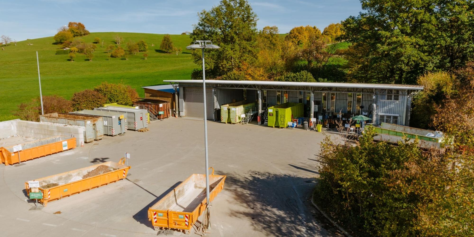 Drohnenaufnahme des Wertstoffzentrums, im Vordergrund drei Bauschuttmulden, im hinteren Teil überdacher Bereich mit weiteren Containern, im Hintergrund Wiese und Bäume.