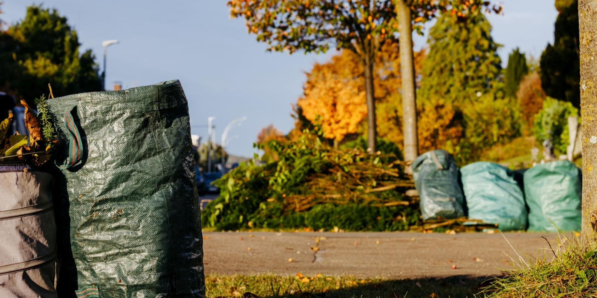 Im Vordergrund links stehen zwei Gartenbags, im Hintergrund liegen mehrere Grüngutbündel neben weiteren Gartenbags.