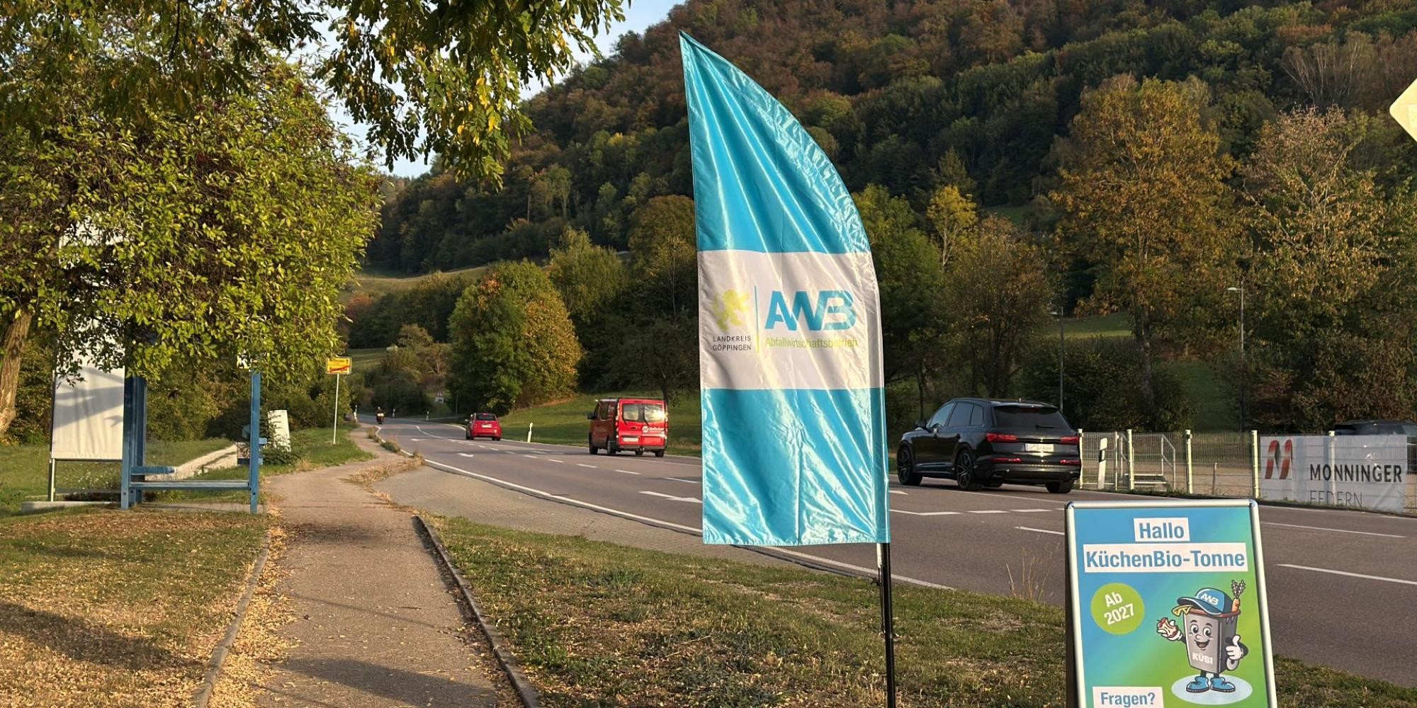 Am Straßenrand steht eine AWB-Beachflag, daneben ein Kundenstopper, auf dem Kübi abgebildet ist, mit der Aufschrift "Hallo KüchenBio-Tonne. Fragen? Hier gibt's Antworten". Auf der Straße fahren Fahrzeuge, im Hintergrund Wald.