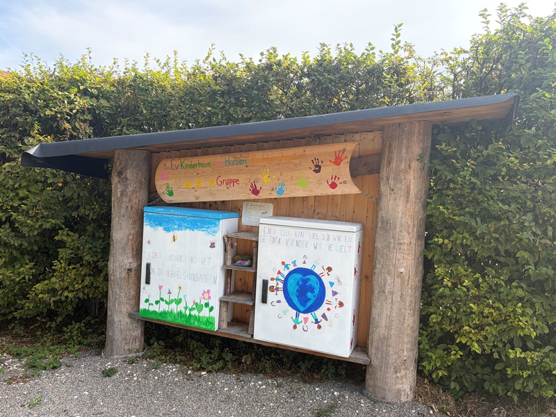 Überdachte hölzerne Bücherstation mit zwei bunt bemalten Schränken, in denen Bücher abgegeben und getauscht werden können. Darüber hängt ein handbemaltes Schild des Evangelischen Kinderhauses Horben in Rechberghausen. Im Hintergrund befindet sich eine dichte grüne Hecke.